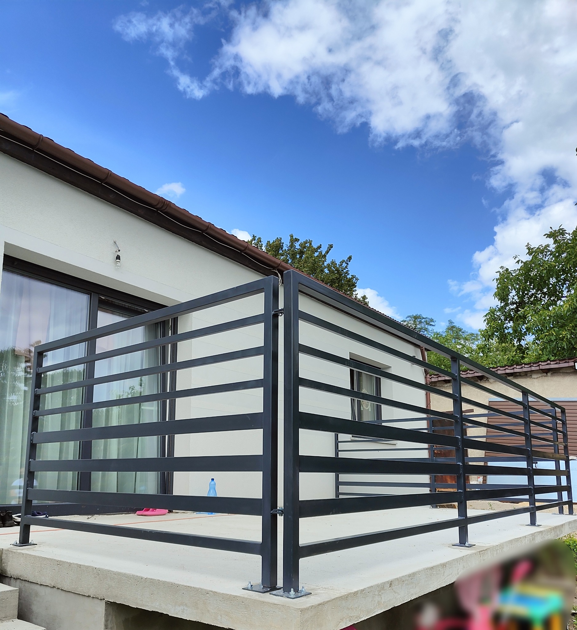 Nowoczesna, czarna balustrada z metalowych profili na betonowym tarasie, z widocznymi detalami mocowania do podłoża oraz elementami otoczenia, takimi jak dom z dużym oknem i błękitne niebo z chmurami.