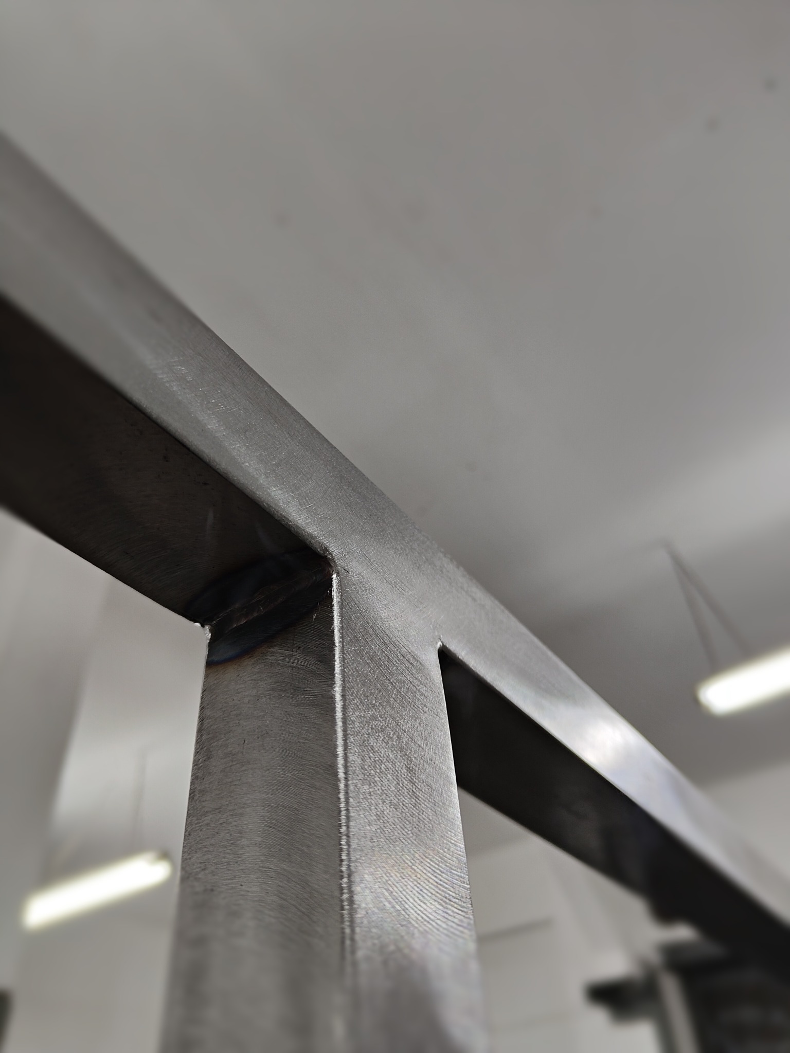 Szczegół spawu na konstrukcji z profili metalowych, widoczne ślady szlifowania, surowy metal w szarym odcieniu, oświetlenie warsztatowe w tle.