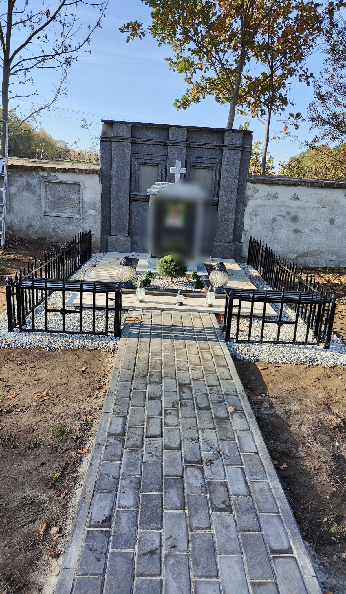 Czarne metalowe ogrodzenie otacza pomnik na cmentarzu, z wybrukowaną ścieżką prowadzącą do grobu.