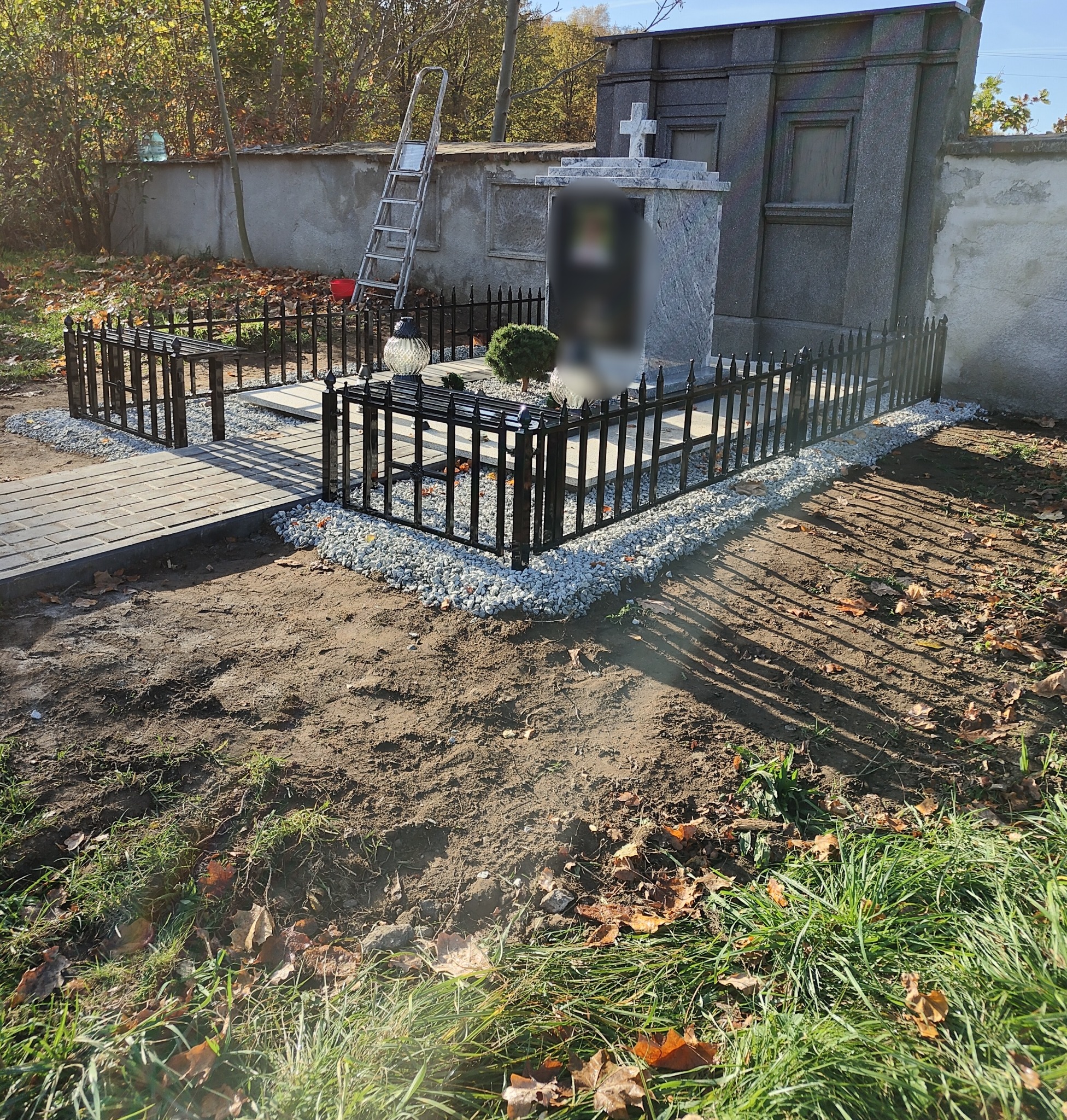 Czarne, metalowe ogrodzenie wokół nagrobka, wykończone drobnym grysem, z brukowanym chodnikiem i drabiną opartą o mur w tle.