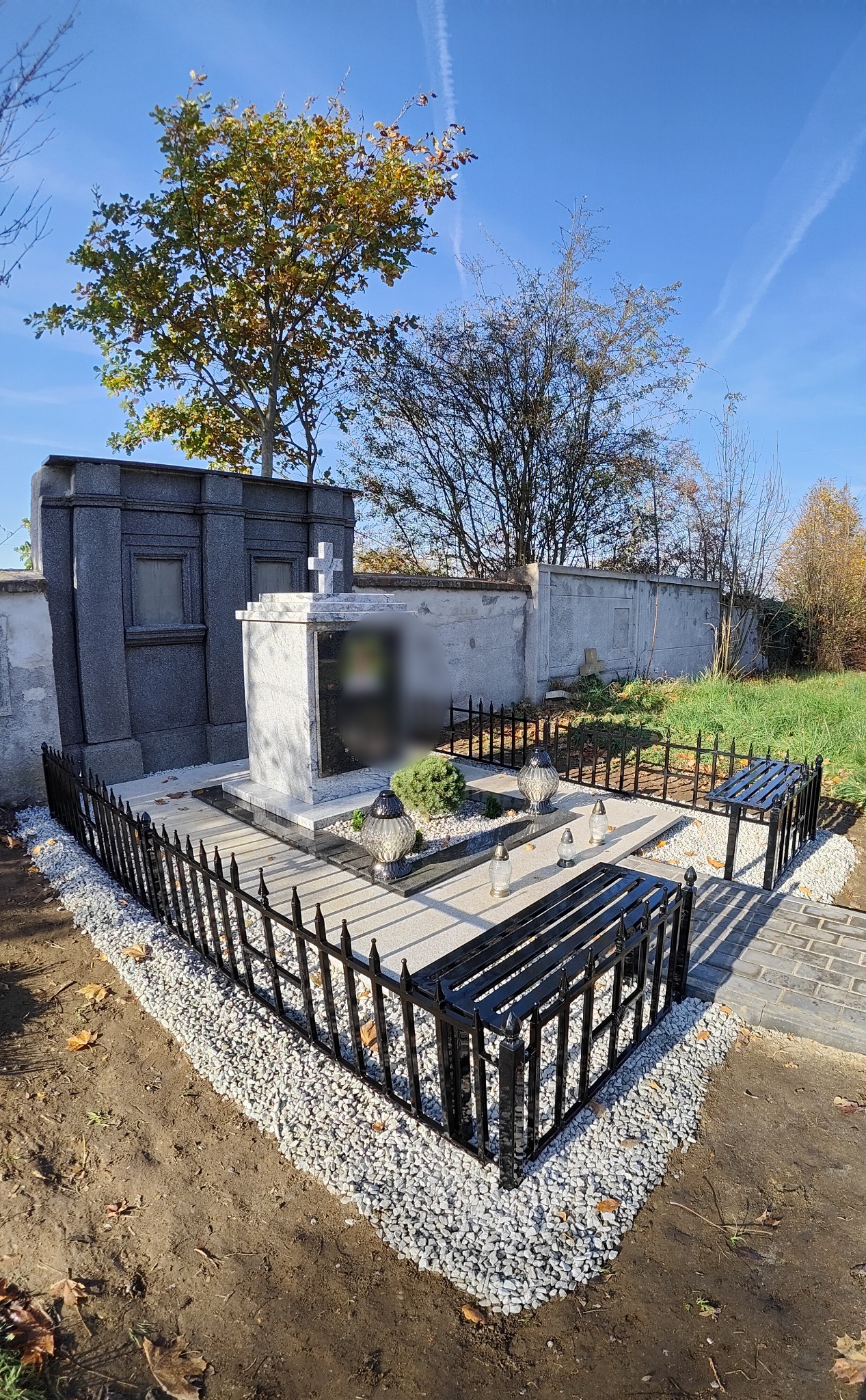 Nagrobek z marmuru otoczony niskim, czarnym ogrodzeniem metalowym z dwoma ławkami, całość wysypana drobnymi, białymi kamieniami, w tle nagrobki i drzewa.