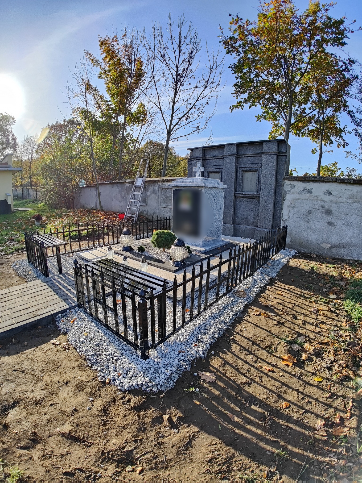 Metalowe ogrodzenie z ławkami otaczające nagrobek na cmentarzu, z drobnymi kamieniami i płytami chodnikowymi.