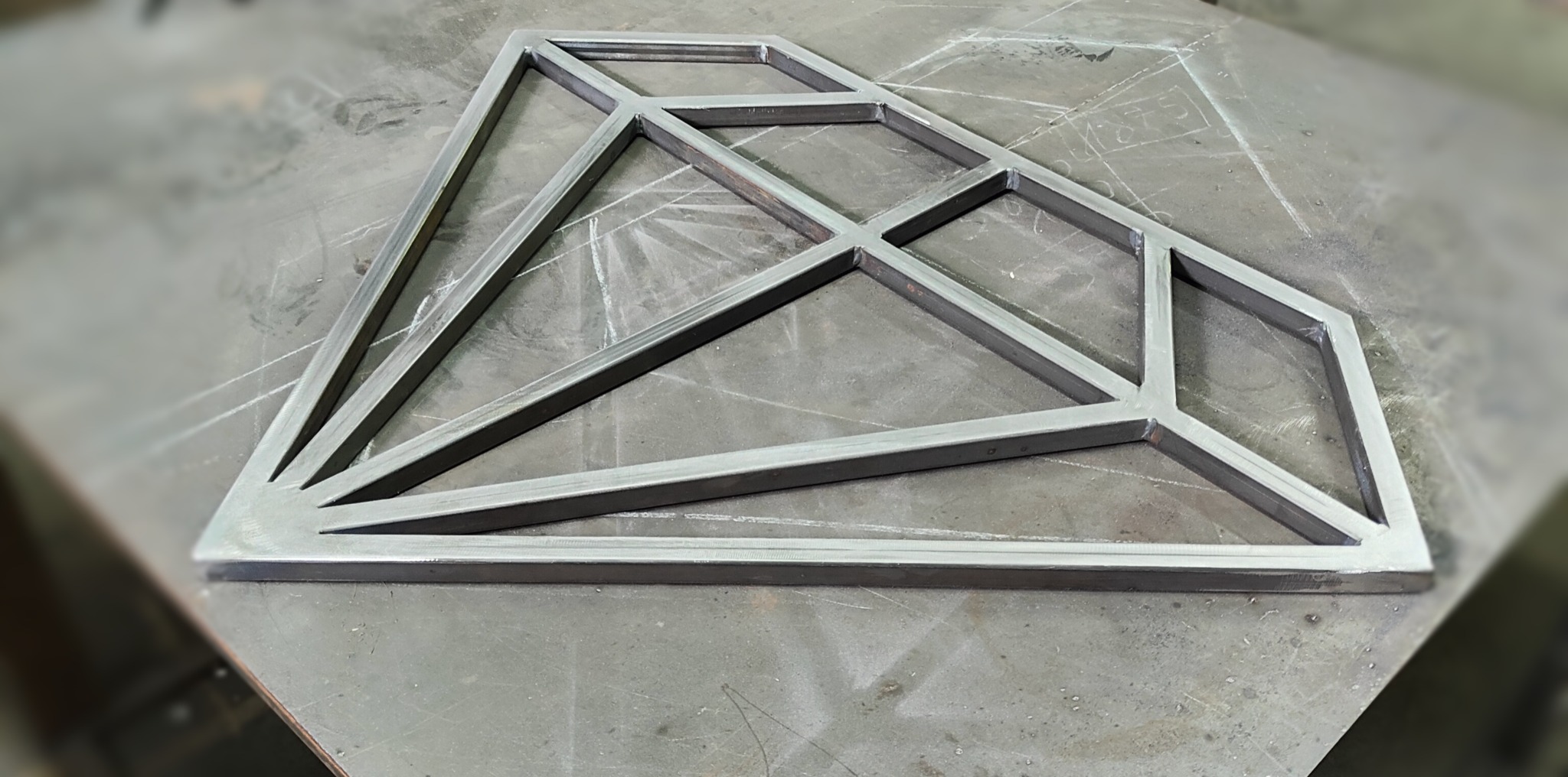 Surowa metalowa konstrukcja w kształcie diamentu, spawana z profili, leżąca na szarym, roboczym stole z widocznymi śladami po cięciu i spawaniu.