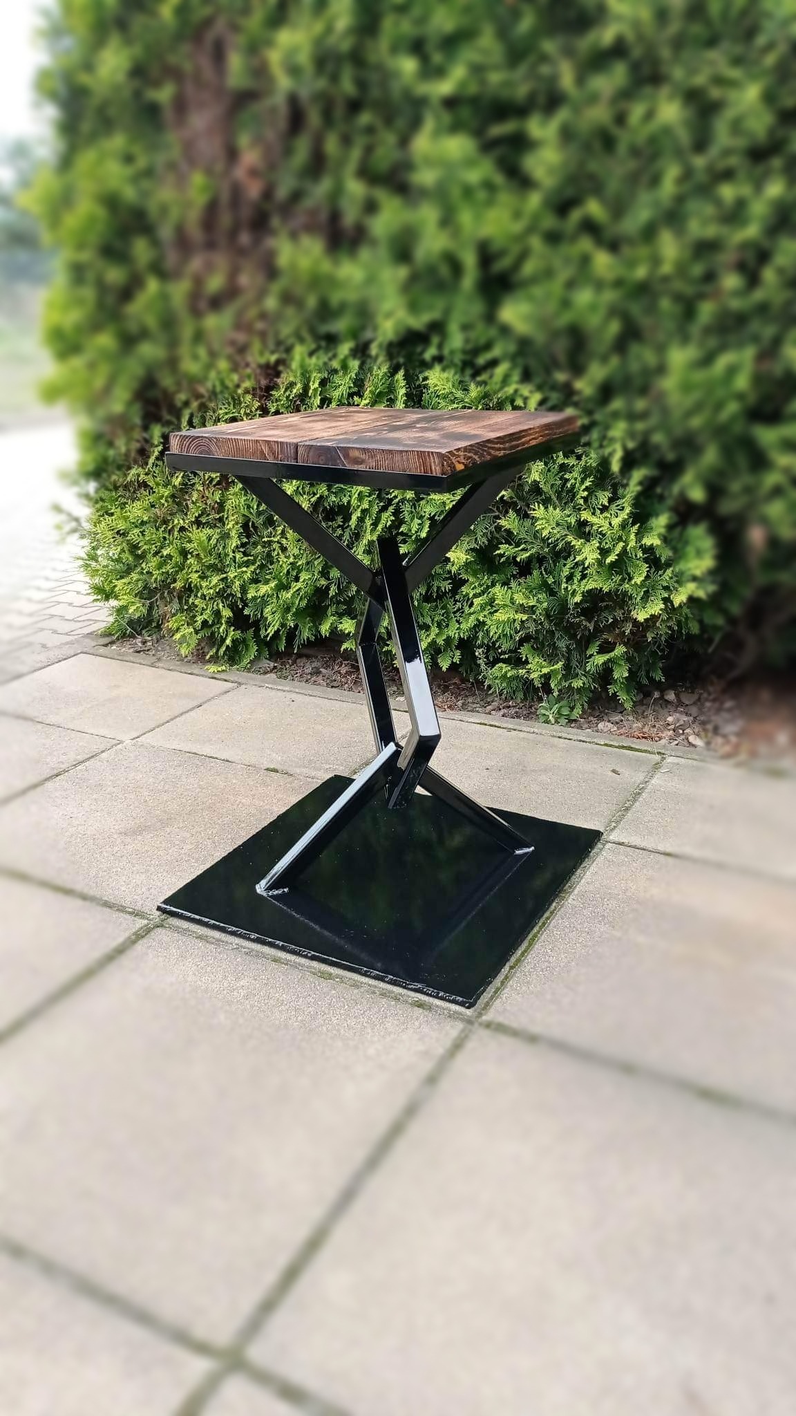 Stolik kawowy z drewnianym blatem i geometryczną podstawą z czarnego metalu, ustawiony na brukowanej powierzchni przed zielonym żywopłotem.