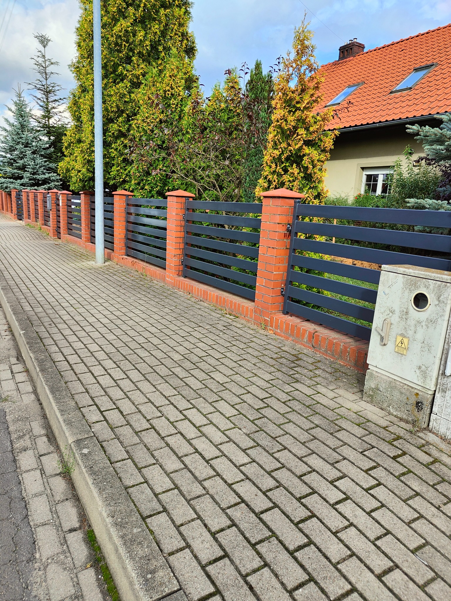 Nowoczesne ogrodzenie posesji z czarnych, poziomych paneli metalowych i słupków z czerwonej cegły, widok z chodnika wyłożonego kostką brukową, w tle dom z czerwoną dachówką i iglaki.