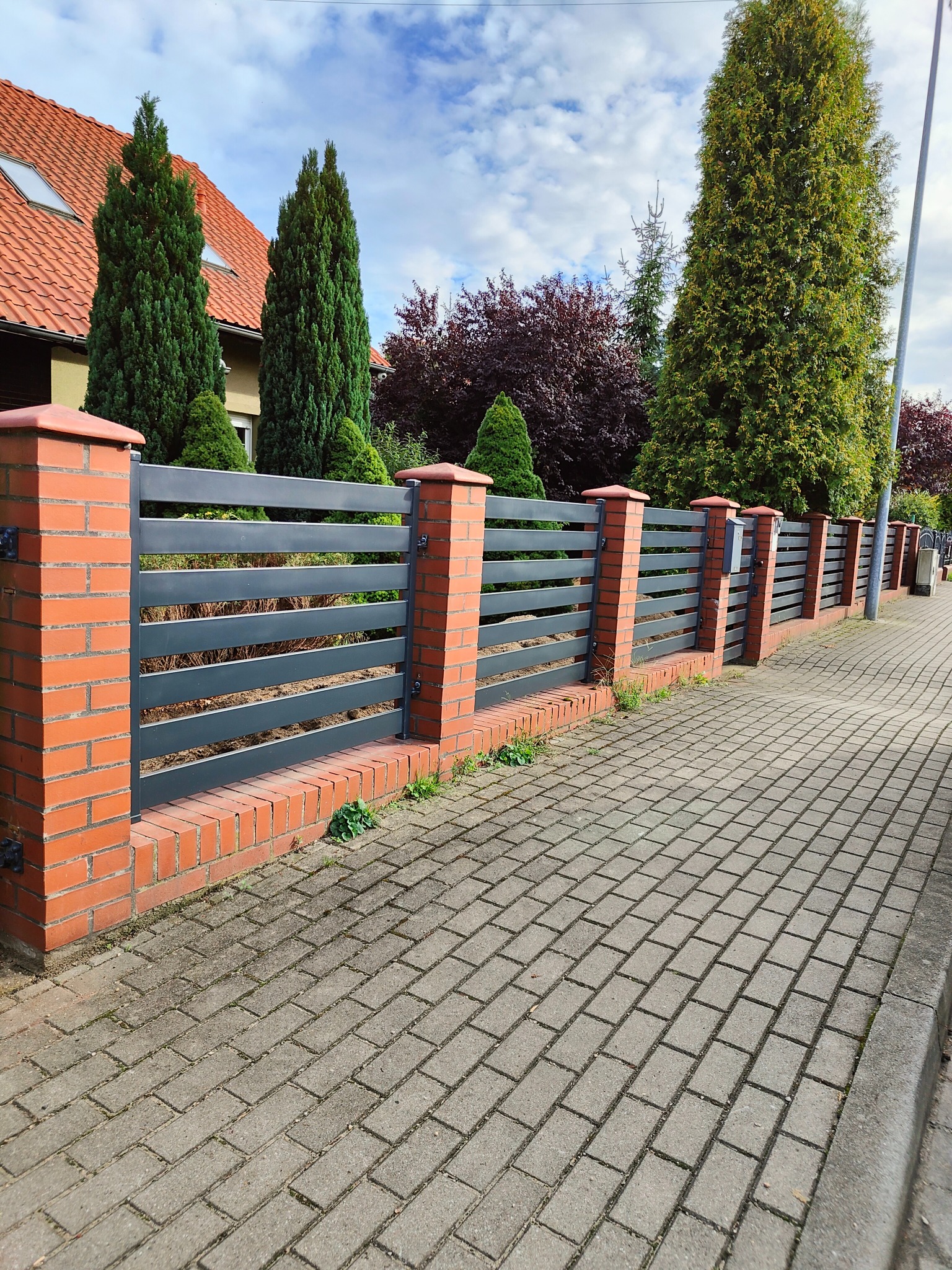 Nowoczesne ogrodzenie metalowe z poziomymi panelami w kolorze grafitowym, osadzone na ceglanych słupkach, widok z perspektywy chodnika z kostki brukowej, w tle dom z czerwoną dachówką i zielone...
