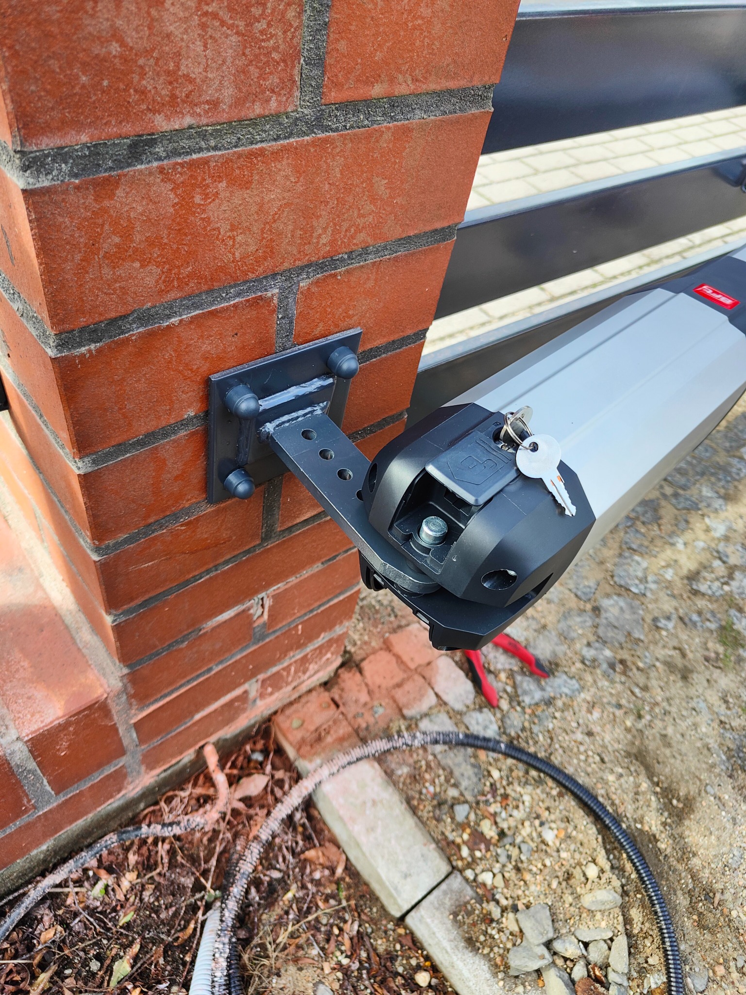 Automatyka bramy wjazdowej zamontowana na ceglanym słupku, widoczny silnik z kluczykiem, mocowanie do słupka z metalu, przewody w ziemi.