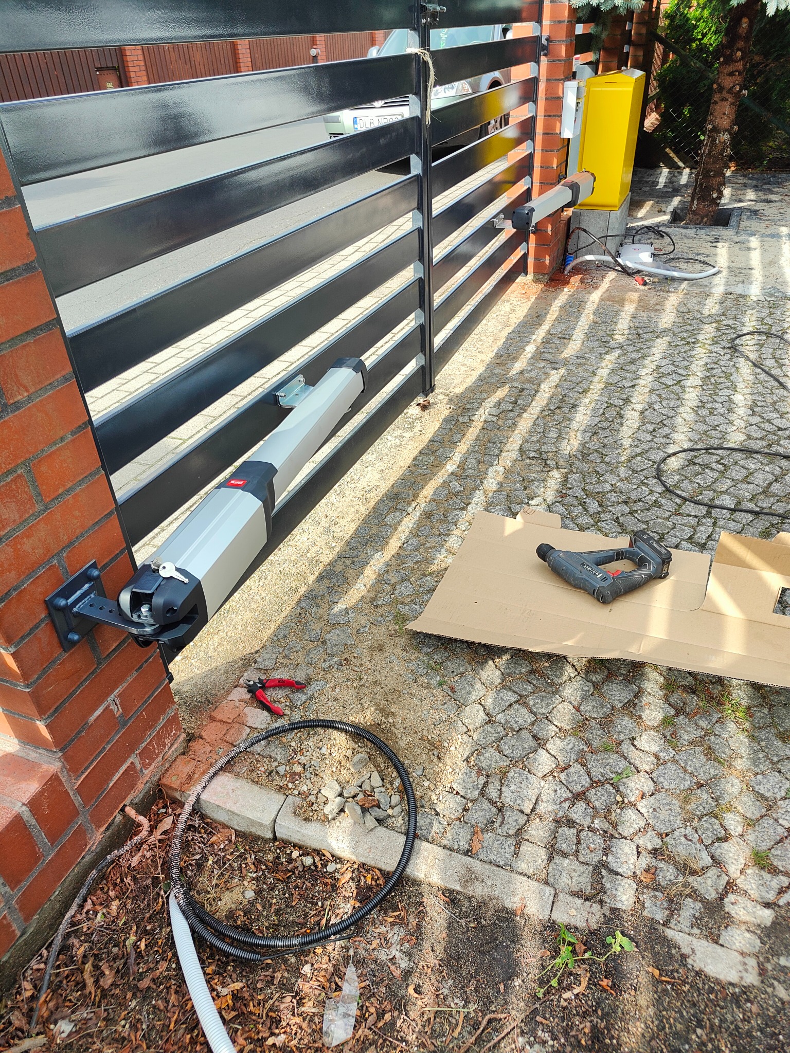 Czarna, pozioma brama wjazdowa z automatycznym systemem otwierania, widoczne siłowniki, skrzynka sterownicza, narzędzia na brukowanej powierzchni, w tle ceglany słupek.