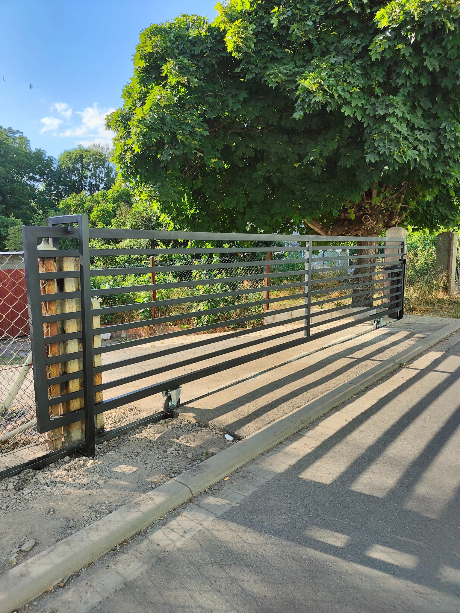 Przesuwna brama wjazdowa z poziomymi profilami, zamontowana na betonowym fundamencie, widoczna od strony ulicy z cieniem rzucanym przez konstrukcję.