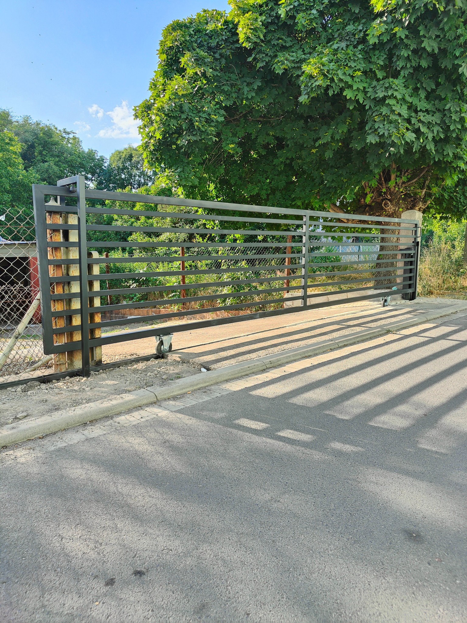 Suwana brama wjazdowa z poziomych metalowych profili, zamontowana na metalowych słupkach, z widocznymi kółkami jezdnymi na tle zieleni i asfaltowej drogi.