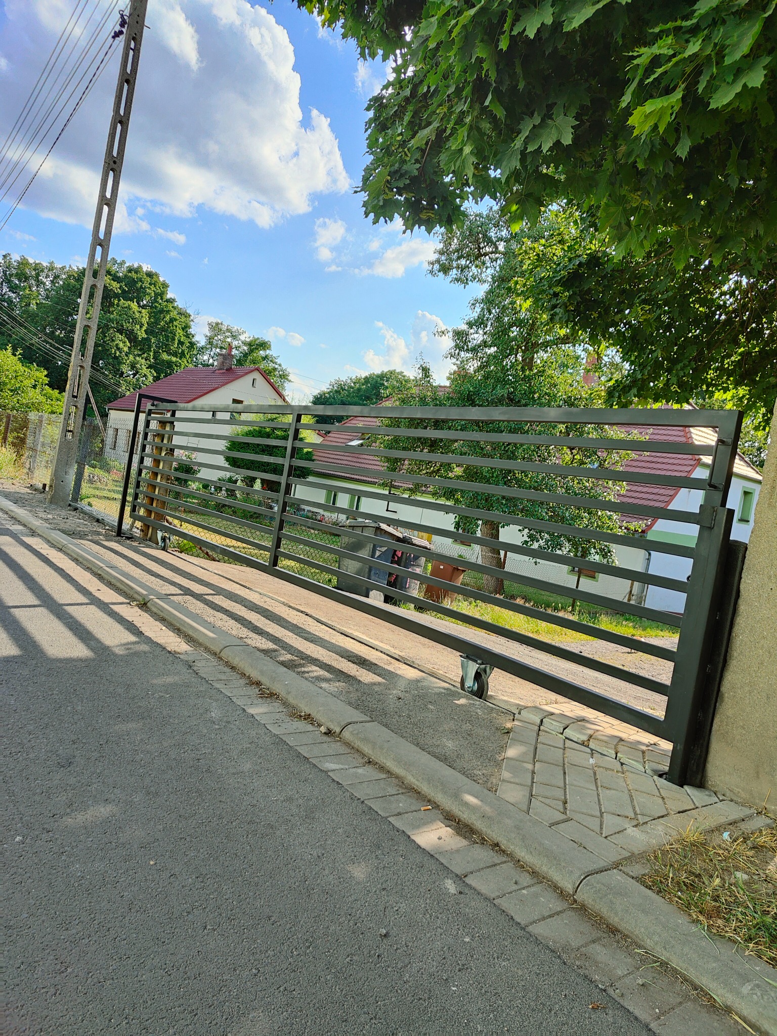 Przesuwna brama wjazdowa z poziomych profili, zamontowana na kółkach, widok z zewnątrz posesji, w tle dom z czerwonym dachem i drzewa.