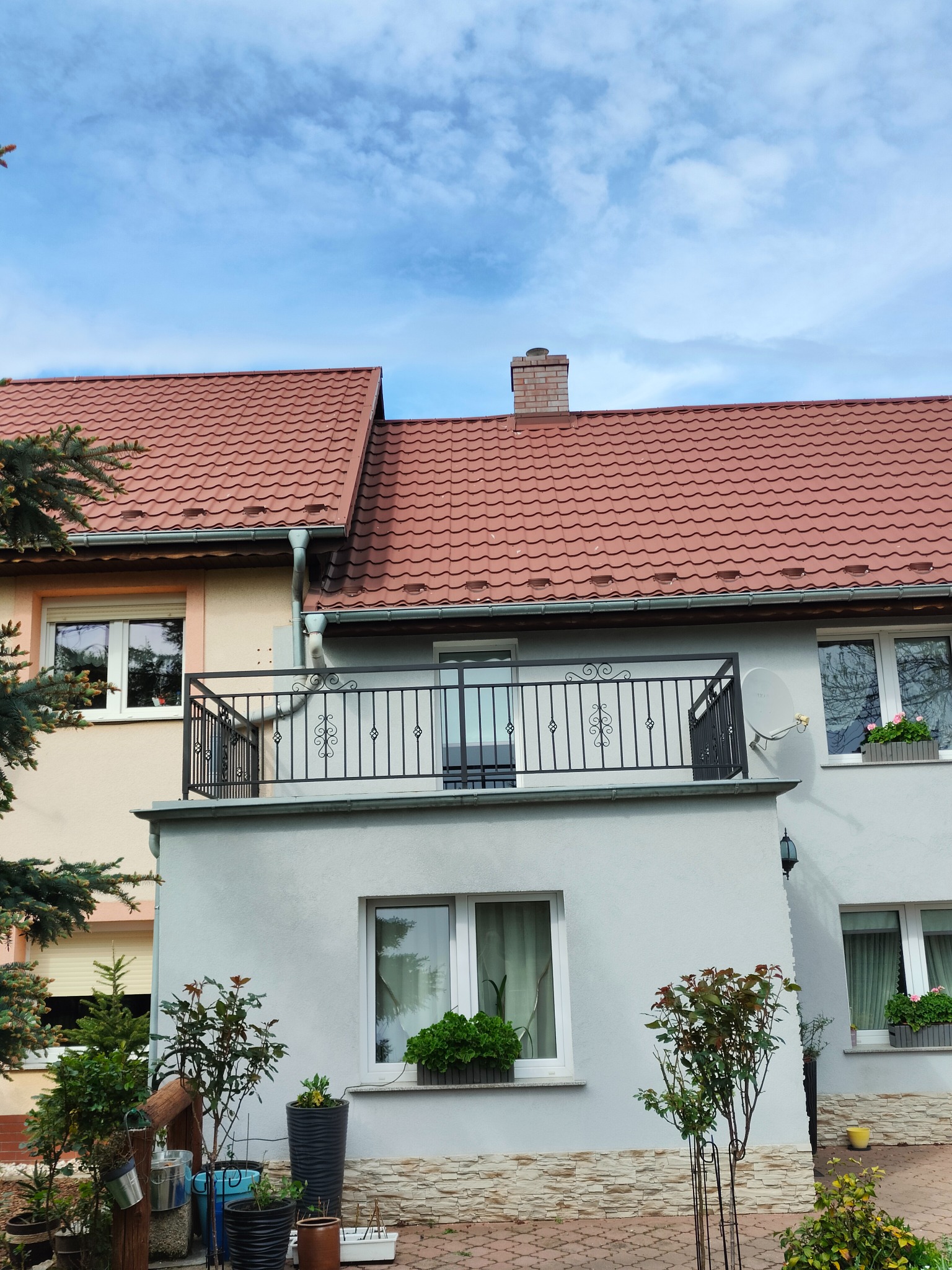 Metalowa balustrada balkonowa z ozdobnymi elementami na tle błękitnego nieba, widok na dom z czerwonym dachem i roślinami w donicach.