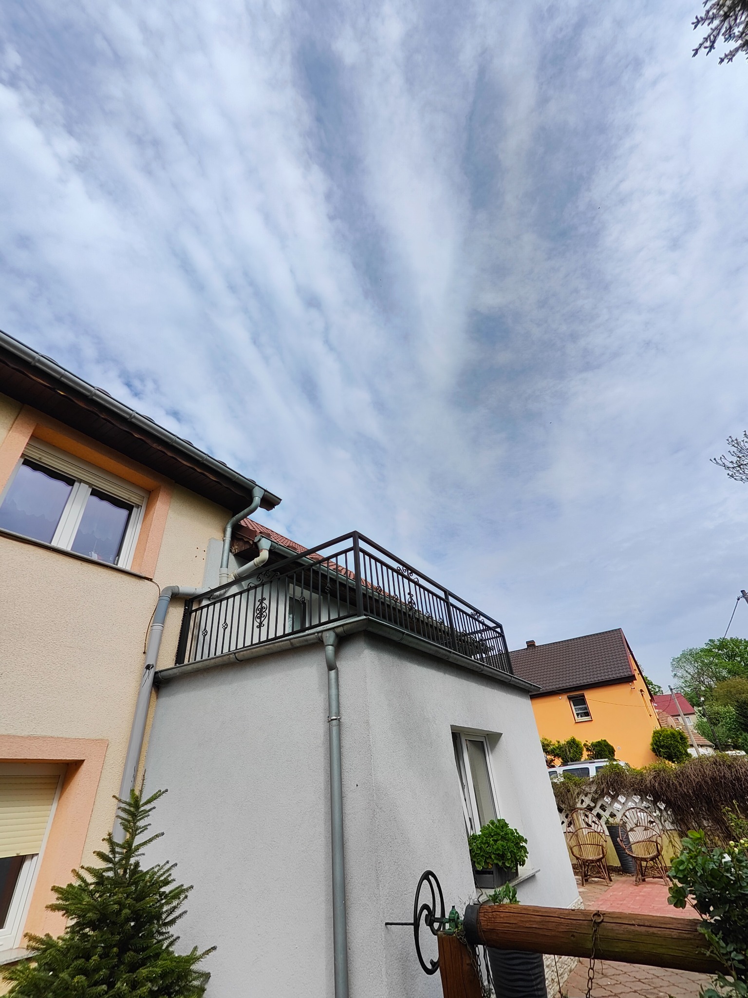 Zewnętrzny balkon z czarną metalową balustradą, widok z dołu na tle pochmurnego nieba, fragment elewacji budynku z oknem i małą choinką.
