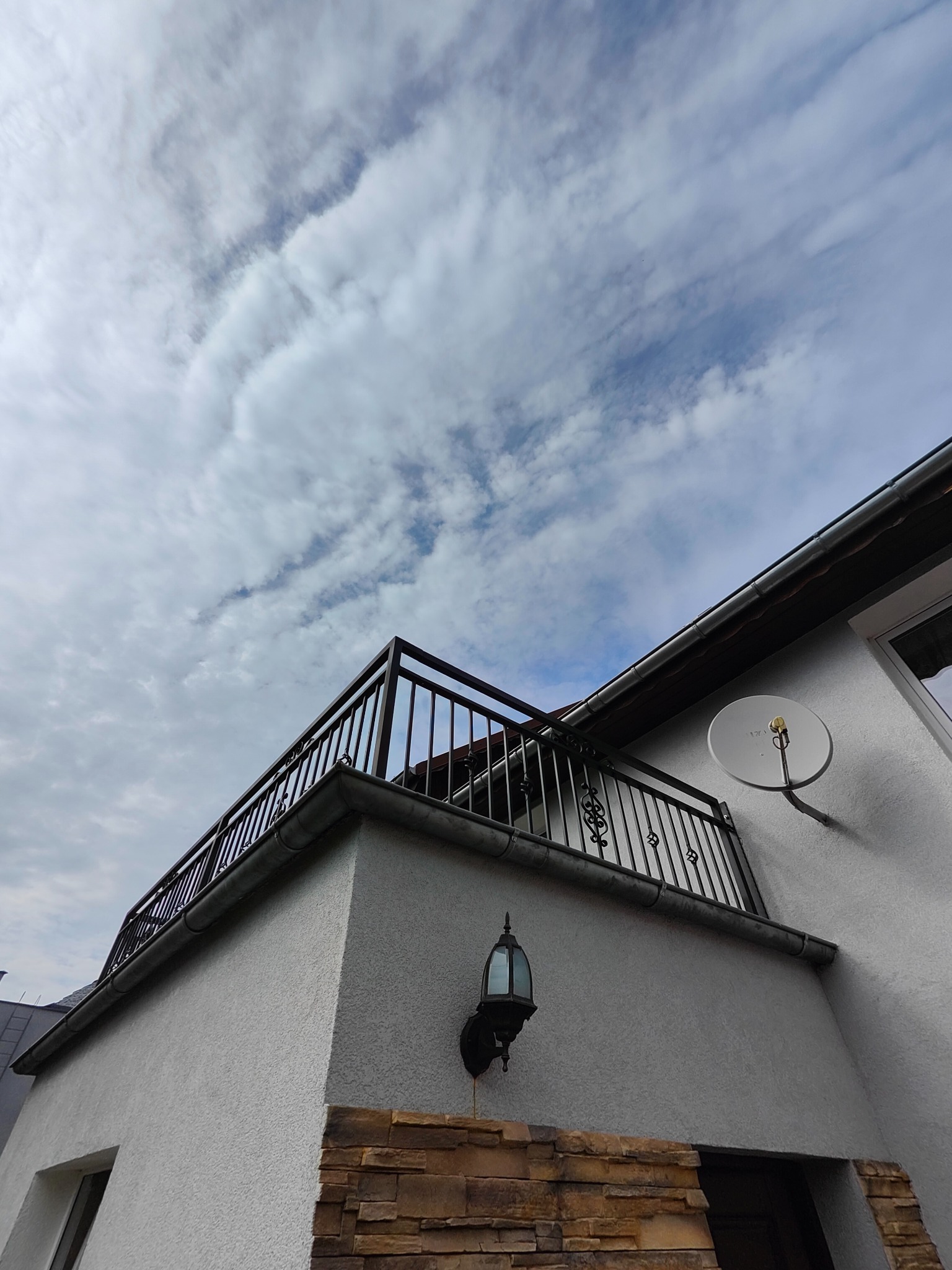 Czarna balustrada balkonowa z metalowymi zdobieniami, zamontowana na białym budynku z widoczną lampą zewnętrzną i anteną satelitarną na tle pochmurnego nieba.