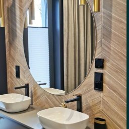 A.S Glass - Nowoczesna łazienka z owalnym lustrem, drewnopodobnymi płytkami ułożonymi w jodełkę, dwoma białymi umywalkami nablatowymi i czarną armaturą, oświetlona złotymi lampami tubami.