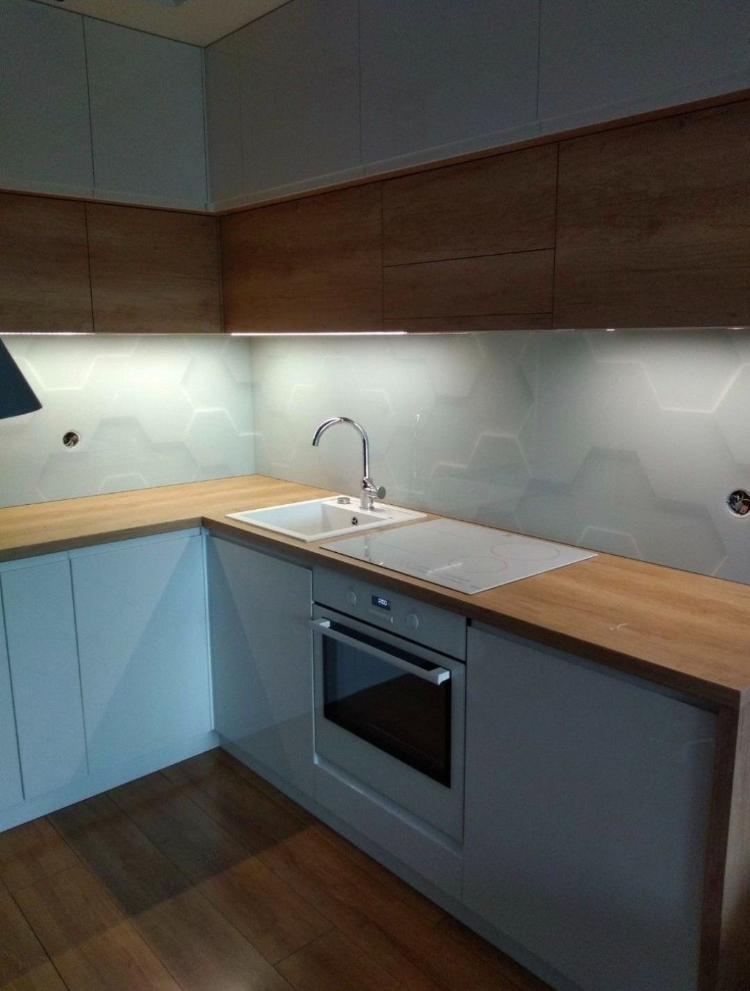 Nowoczesna kuchnia z białymi szafkami, drewnianymi blatami i heksagonalnymi płytkami szklanymi na ścianie, oświetlenie LED pod szafkami górnymi.