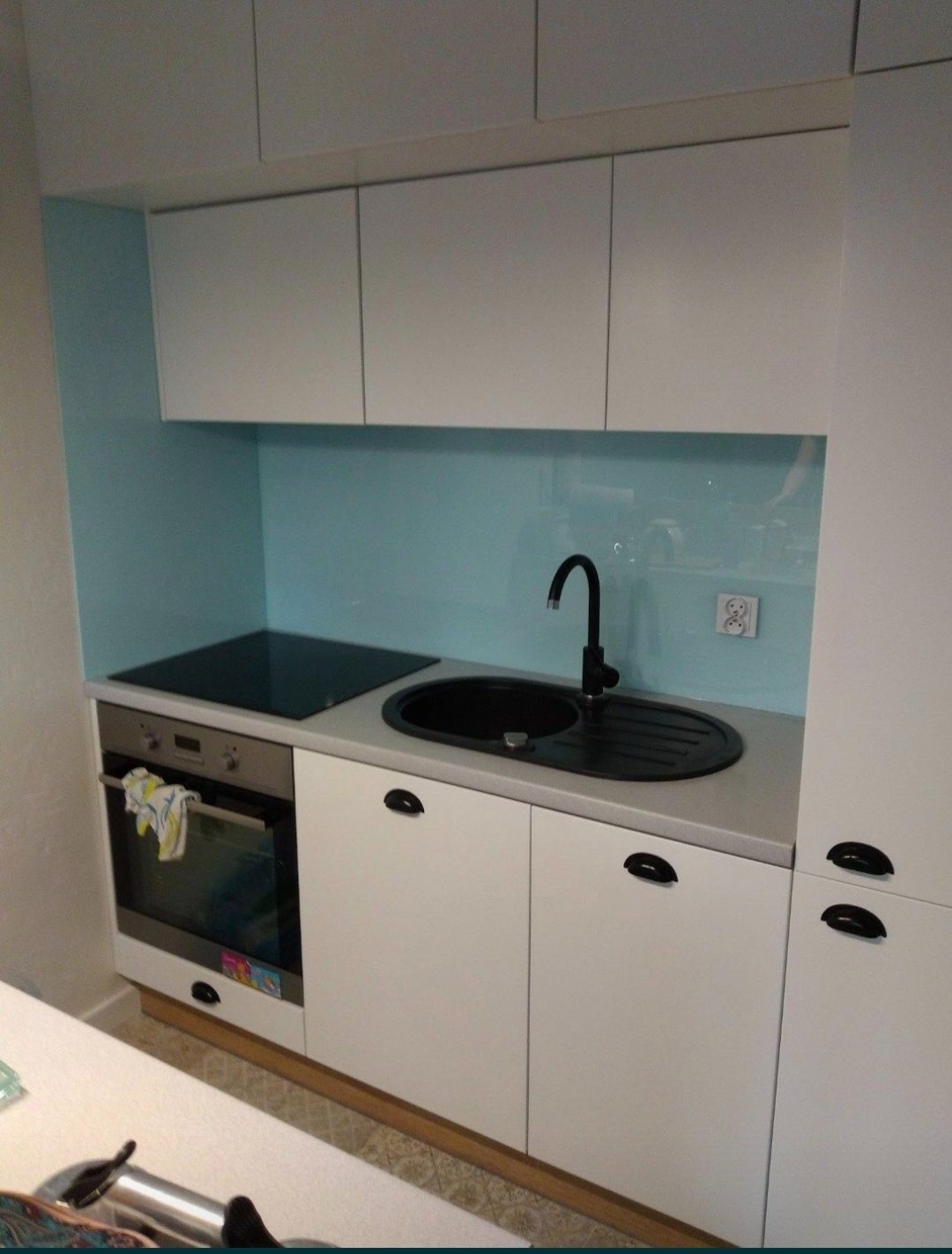 Biała zabudowa kuchenna z turkusowym panelem szklanym nad blatem, czarny zlew i płyta indukcyjna, piekarnik z ręcznikiem w środku.