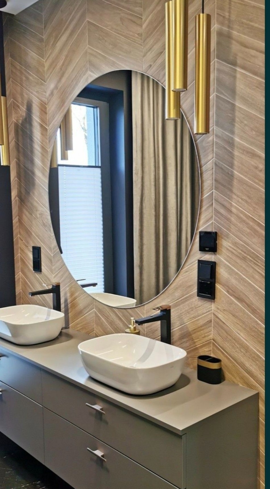 Nowoczesna łazienka z owalnym lustrem, drewnopodobnymi płytkami ułożonymi w jodełkę, dwoma białymi umywalkami nablatowymi i czarną armaturą, oświetlona złotymi lampami tubami.