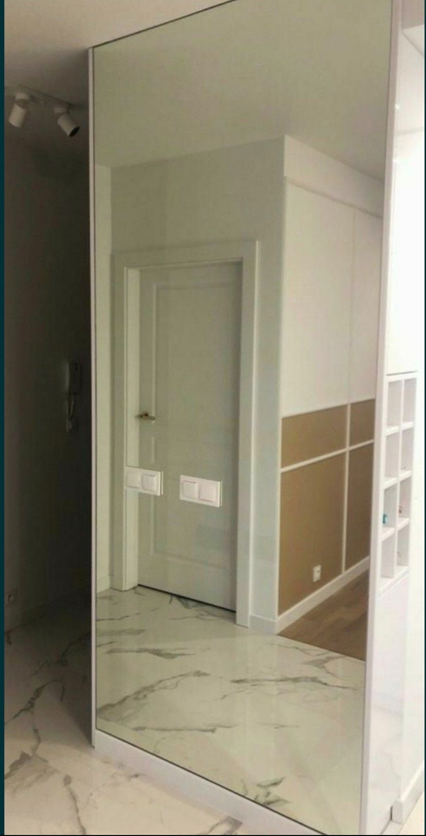 Duże lustro w ramie na ścianie w przedpokoju, odbijające drzwi, białe szafy i marmurową podłogę.