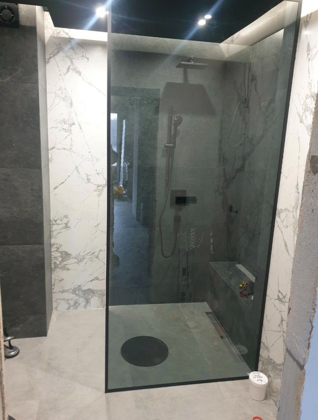 Nowoczesna kabina prysznicowa ze szklaną ścianą, czarną armaturą i odpływem liniowym w posadzce, widoczne niedokończone wykończenia i narzędzia.