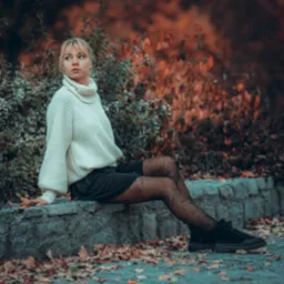 Portret kobiety w białym swetrze i wzorzystych rajstopach, siedzącej na kamiennym murku w jesiennym otoczeniu.