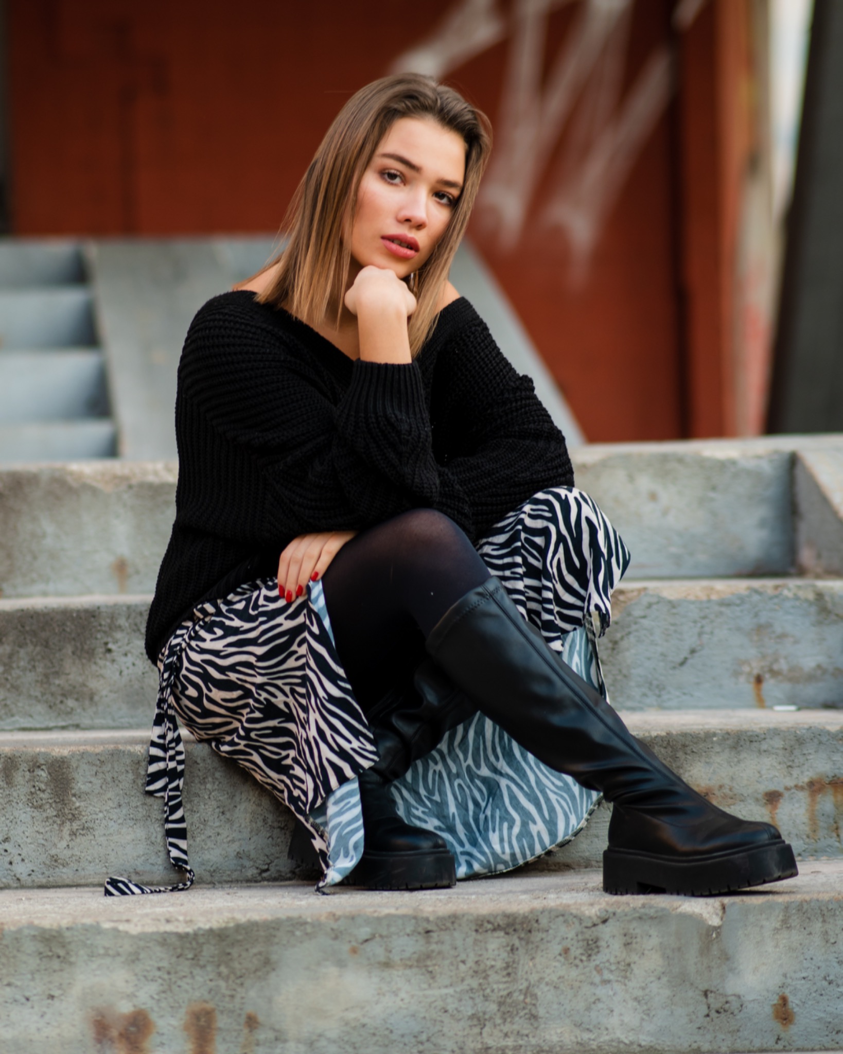 Portret kobiety siedzącej na betonowych schodach, ubrana w czarny sweter, spódnicę w zebrę i wysokie, czarne buty na platformie. Czerwone paznokcie, neutralne tło z graffiti.