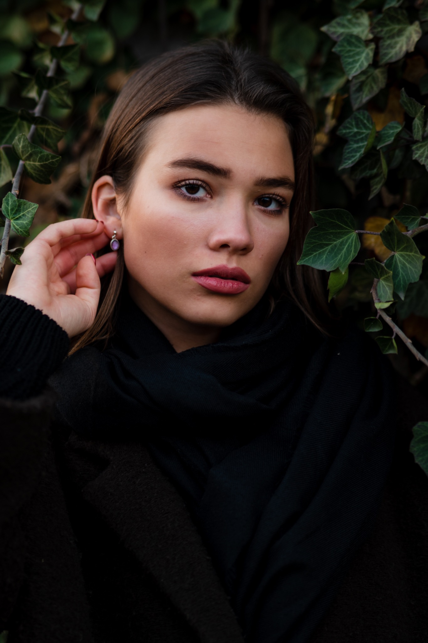 Portret młodej kobiety z kolczykiem na tle zielonego bluszczu, ubrana w ciemny płaszcz i szal.