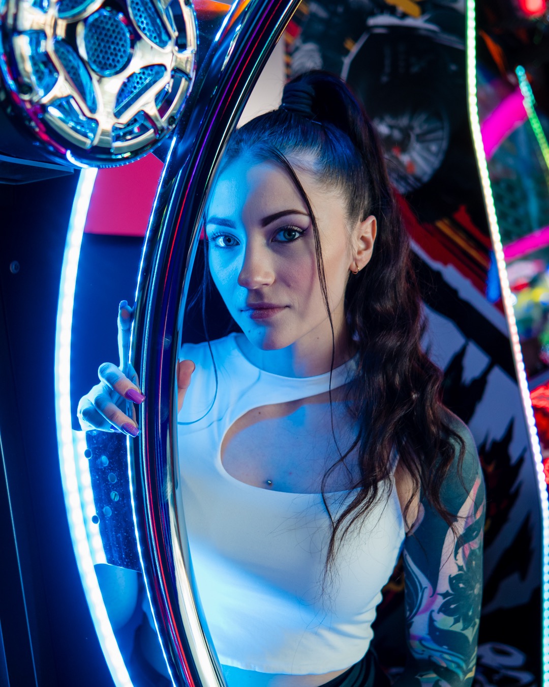 Portret młodej kobiety z ciemnymi włosami w kucyku, w białym topie z wycięciem, opierającej się o neonową konstrukcję w chłodnym, niebiesko-różowym oświetleniu, z tatuażem na ramieniu.