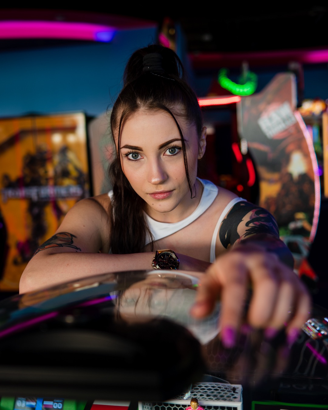 Portret młodej kobiety z tatuażami, opierającej się o automat do gier w salonie gier, z neonowym oświetleniem w tle.