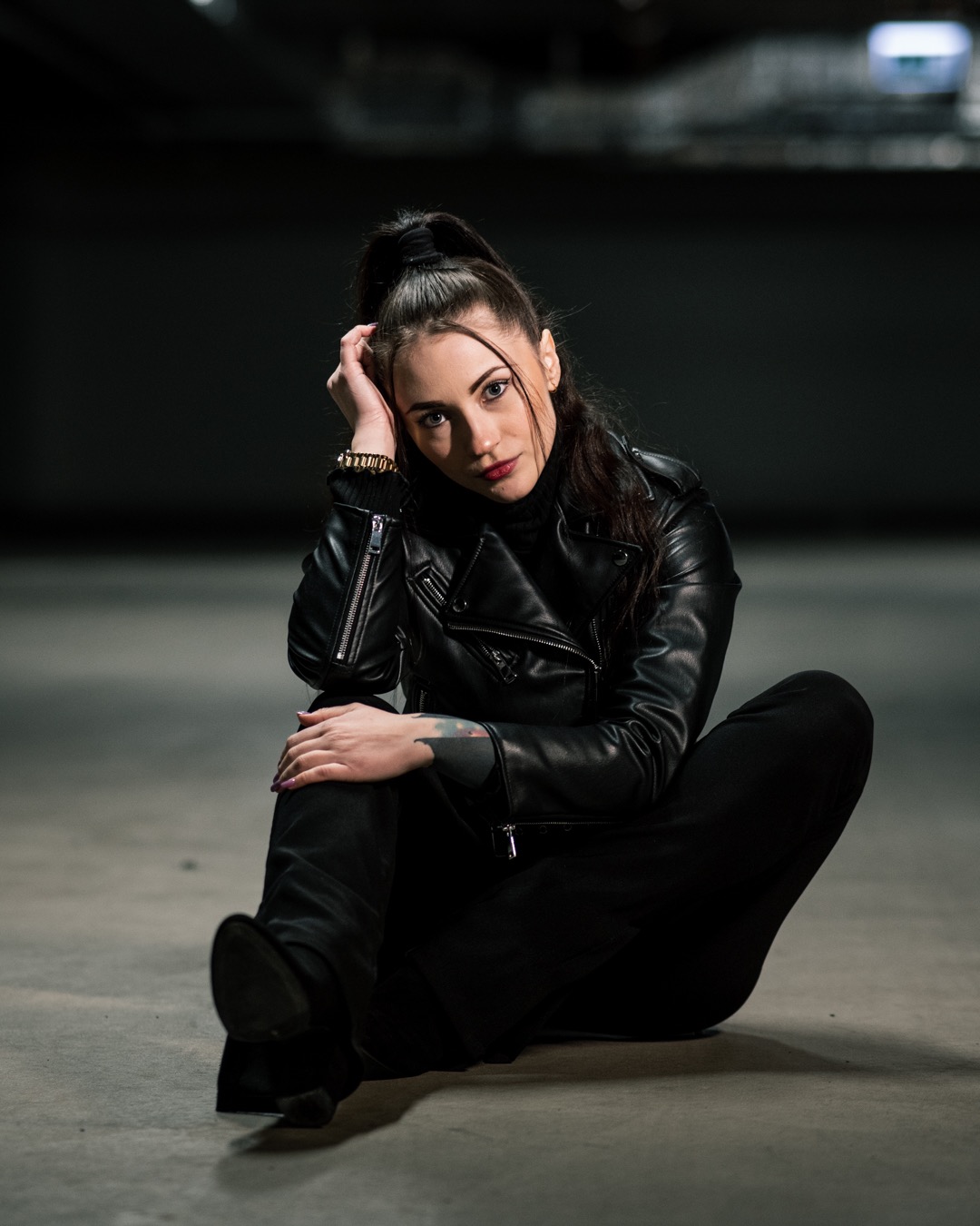 Portret kobiety siedzącej na betonowej podłodze w czarnej skórzanej kurtce i spodniach, z włosami upiętymi w wysoki kucyk i złotą bransoletką na nadgarstku, spoglądającej w obiektyw.