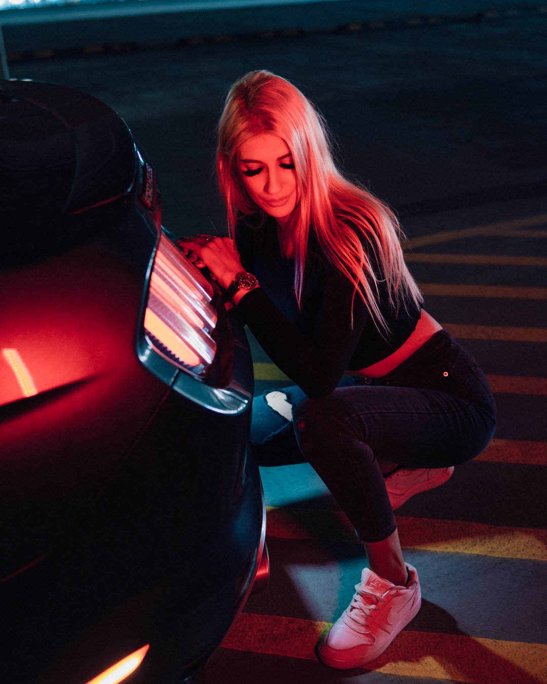 Blondynka w czarnym topie i dżinsach kuca obok czarnego samochodu na parkingu, czerwone światło odbija się od karoserii i twarzy, widoczny zegarek na nadgarstku.