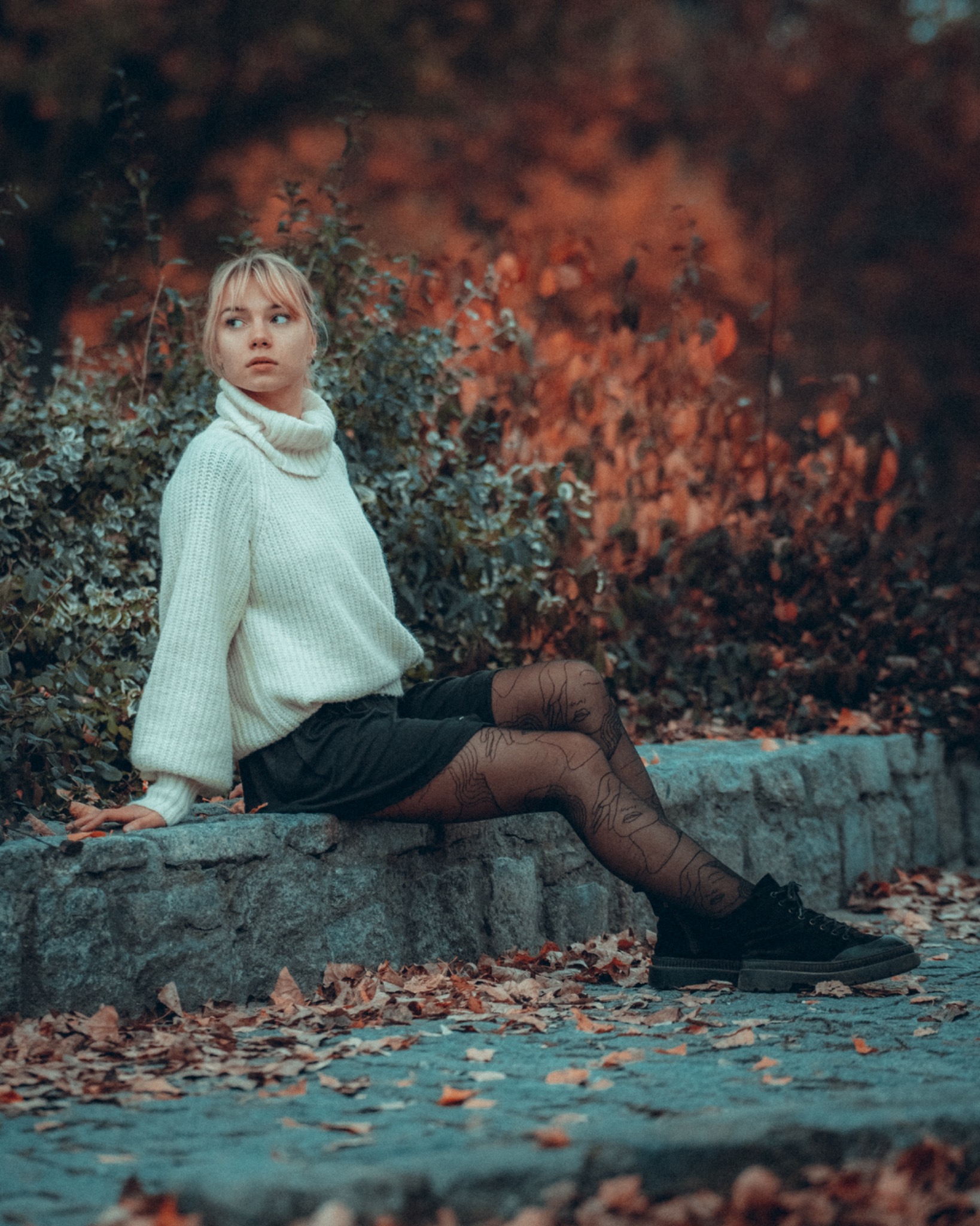 Portret kobiety w białym swetrze i wzorzystych rajstopach, siedzącej na kamiennym murku w jesiennym otoczeniu.