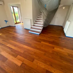 Wycyklinowany, lakierowany parkiet w salonie z zabezpieczonymi taśmą ścianami i schodami pod folią ochronną. Widoczne naturalne usłojenie drewna.