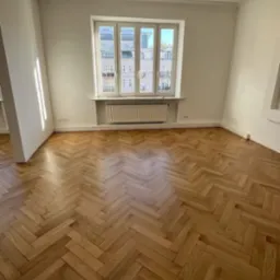 Jasny parkiet ułożony w jodełkę w przestronnym pokoju z oknem. Widoczny grzejnik pod oknem i fragment kolumny. Naturalne światło podkreśla strukturę drewna.