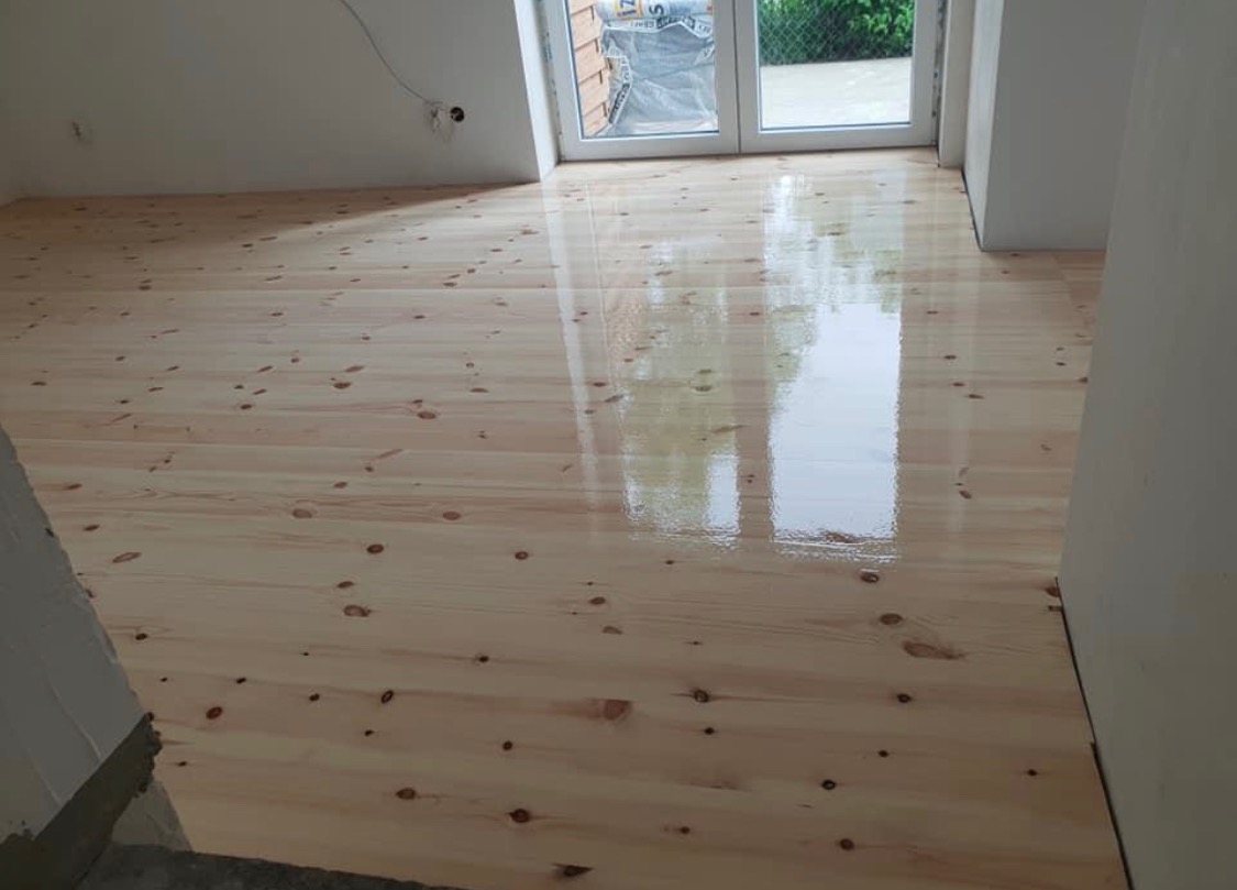 Świeżo położona, błyszcząca podłoga z jasnego drewna z widocznymi sękami w pustym pokoju, odbijająca światło z okna balkonowego.