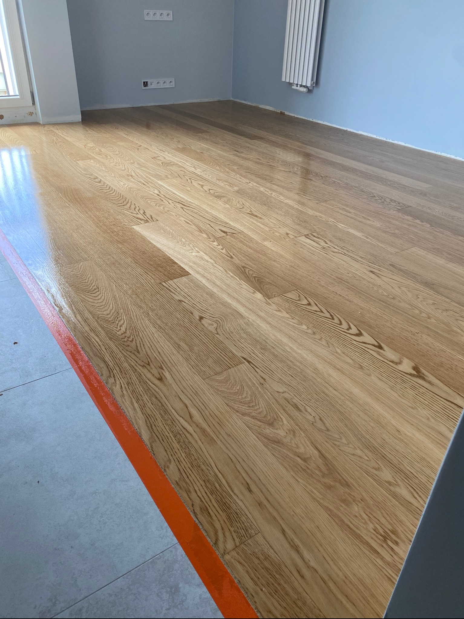 Świeżo polakierowany parkiet dębowy, zabezpieczony taśmą malarską przy przejściu do płytek, widoczne gniazdka elektryczne na ścianie.