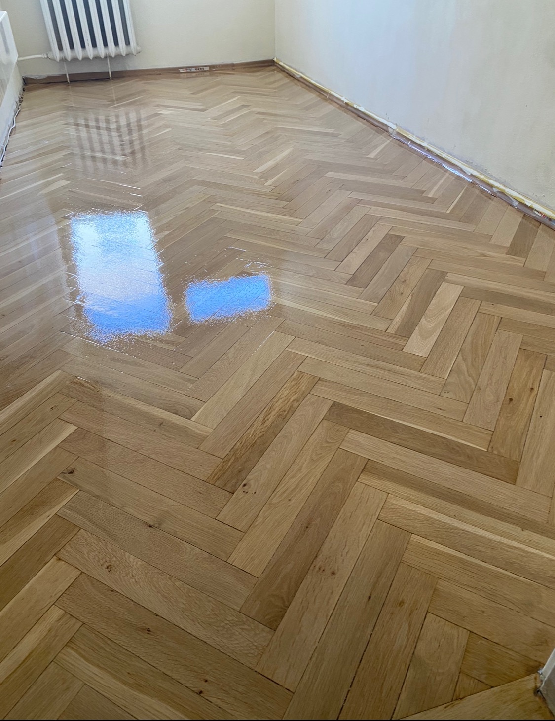 Świeżo polakierowany parkiet ułożony w jodełkę, z widocznym odbiciem okna, listwy przypodłogowe zabezpieczone folią.