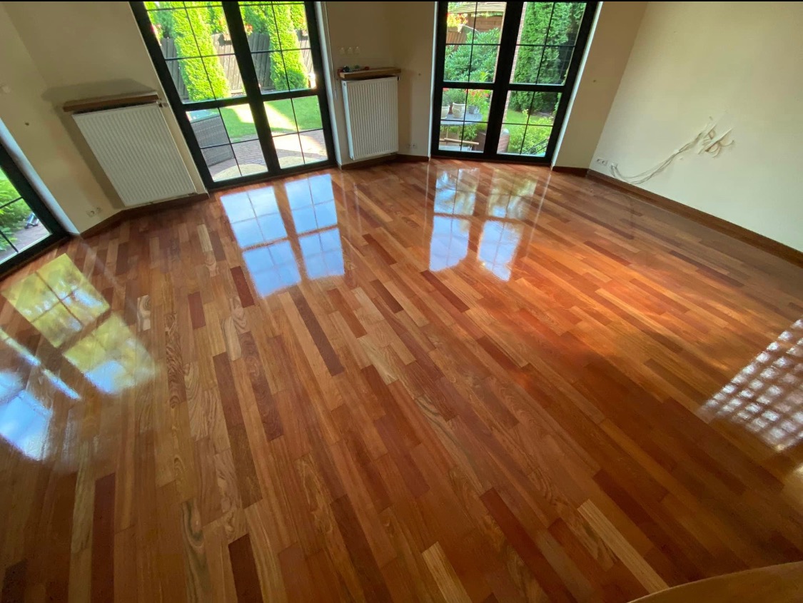 Błyszcząca, świeżo położona parkietowa podłoga w pokoju z dużymi oknami z widokiem na zieleń, odbijającymi się w powierzchni podłogi.