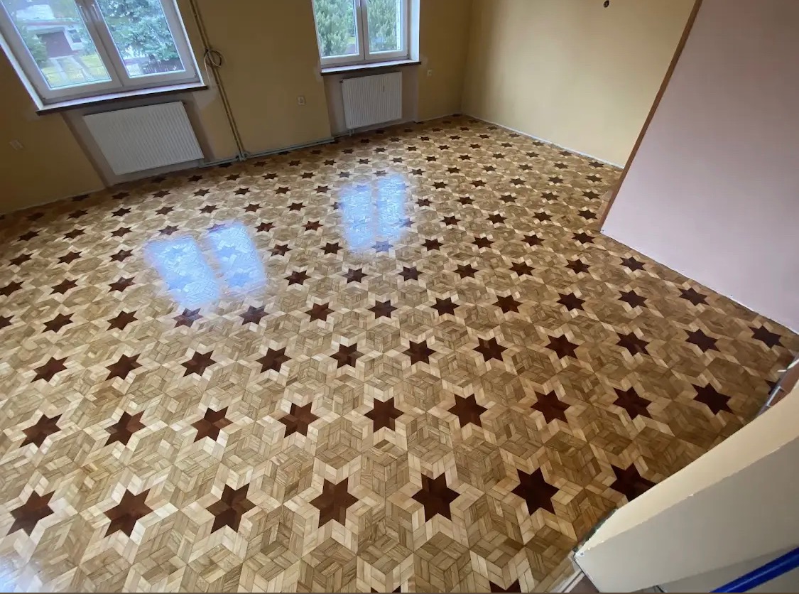 Wykończona podłoga z parkietu ułożonego we wzór gwiazdy na jasnym tle, widoczne dwa okna z grzejnikami pod spodem, ściany w kolorze beżowym.