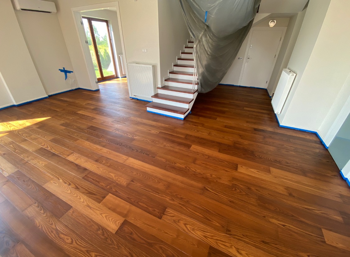 Świeżo polakierowana, brązowa podłoga z desek w jasnym pomieszczeniu, zabezpieczona niebieską taśmą malarską przy ścianach i schodach, które są częściowo zakryte folią ochronną.