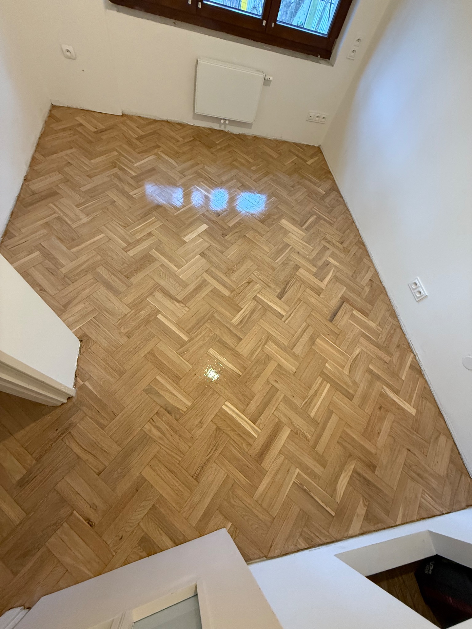 Ułożony parkiet w jodełkę w jasnym pomieszczeniu, widoczne gniazdka elektryczne na ścianie i okno w górnej części kadru, na podłodze widoczne refleksy świetlne.