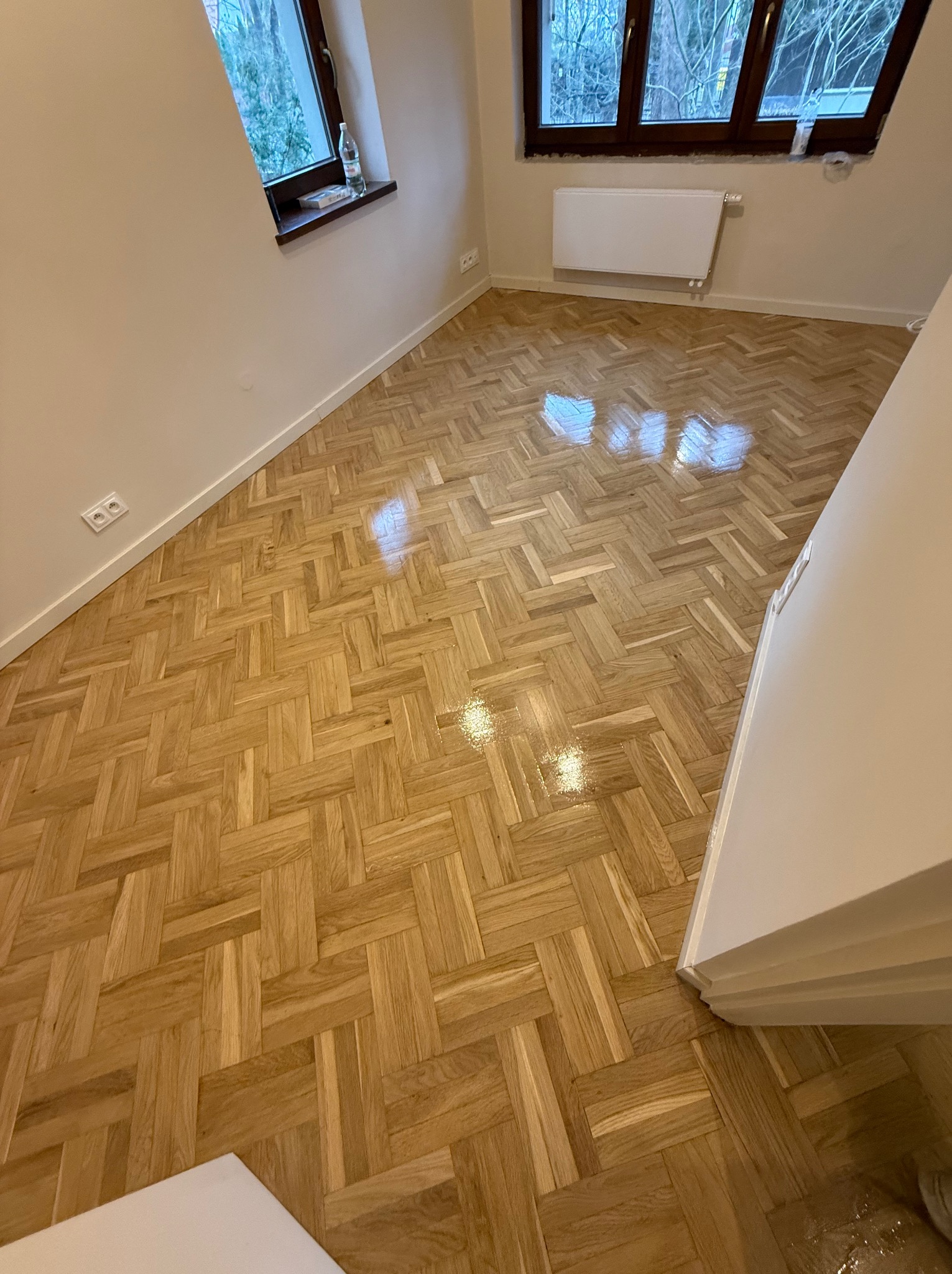 Nowo ułożony parkiet w jodełkę, pokryty błyszczącym lakierem, widoczne odbicia światła, okno z widokiem na drzewa w tle.