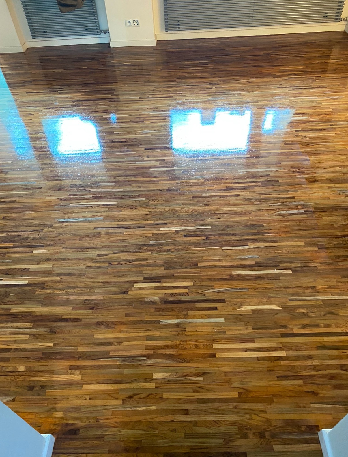 Świeżo polakierowany, błyszczący parkiet z egzotycznego drewna, widoczne odbicia światła z okien, ułożony w regularny wzór.