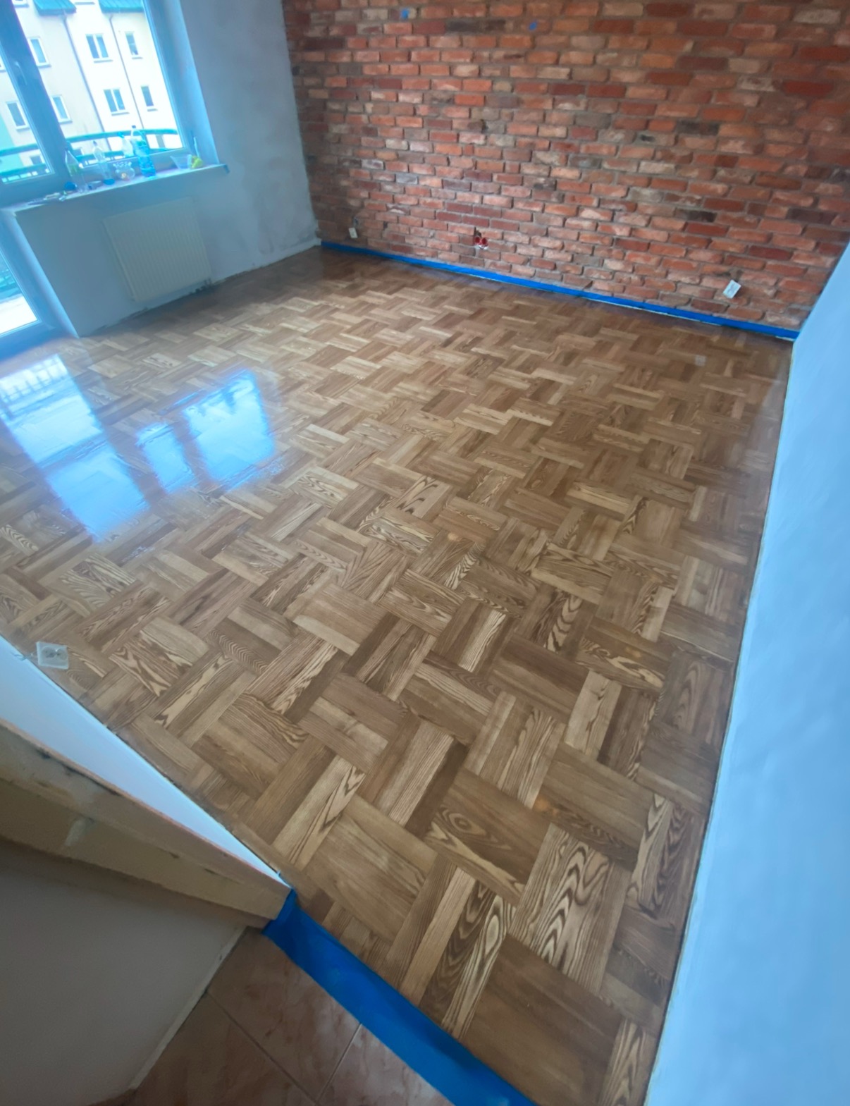 Ułożony parkiet w jodełkę francuską, zabezpieczony niebieską taśmą malarską przy ścianach z surowej cegły i białych ścianach, z widocznym odbiciem światła z okna.