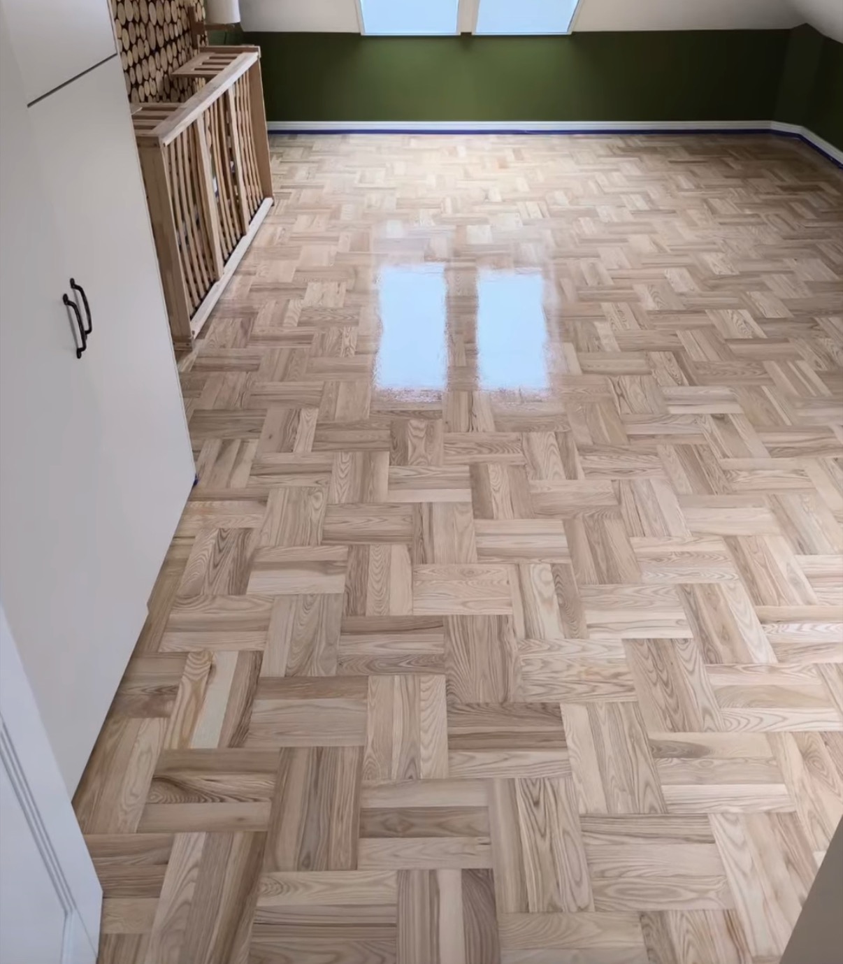 Parkiet ułożony w jodełkę francuską, świeżo po lakierowaniu, z widocznym odbiciem okna w połyskującej powierzchni, w tle ciemnozielona ściana z niebieskim paskiem przy podłodze.