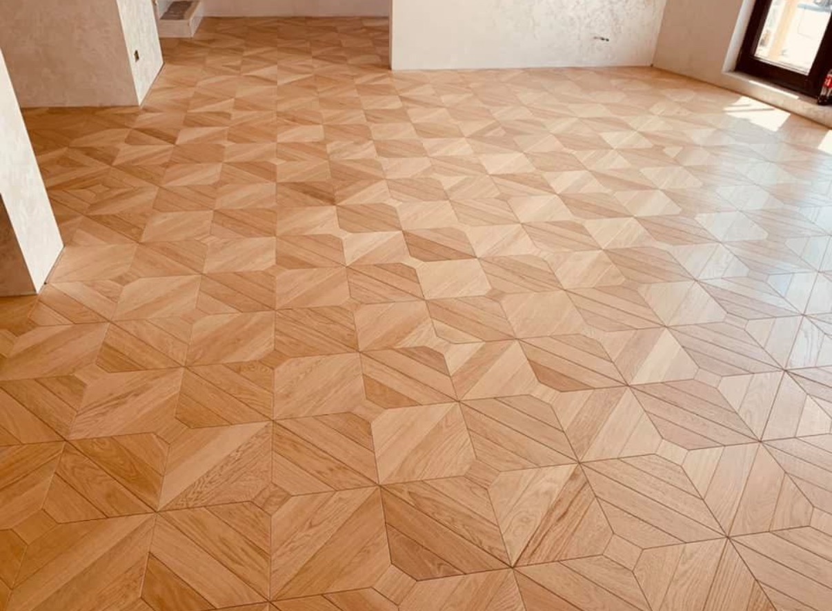 Nowo ułożony parkiet z jasnego drewna w geometryczny wzór gwiazd i kwadratów, widok z góry, z fragmentem ściany i okna w tle.