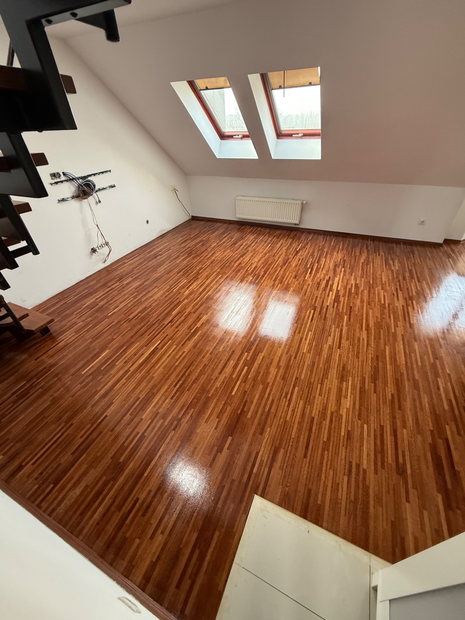Wnętrze poddasza z nowo ułożonym parkietem w odcieniach brązu, widoczne dwa okna dachowe, grzejnik i fragment schodów z czarną balustradą.