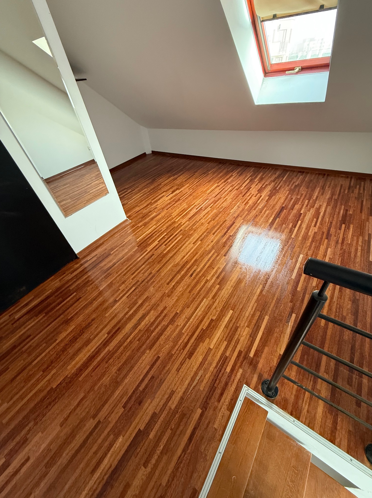 Błyszcząca, świeżo polakierowana podłoga z parkietu w skosach poddasza, widoczny fragment schodów i okno dachowe.