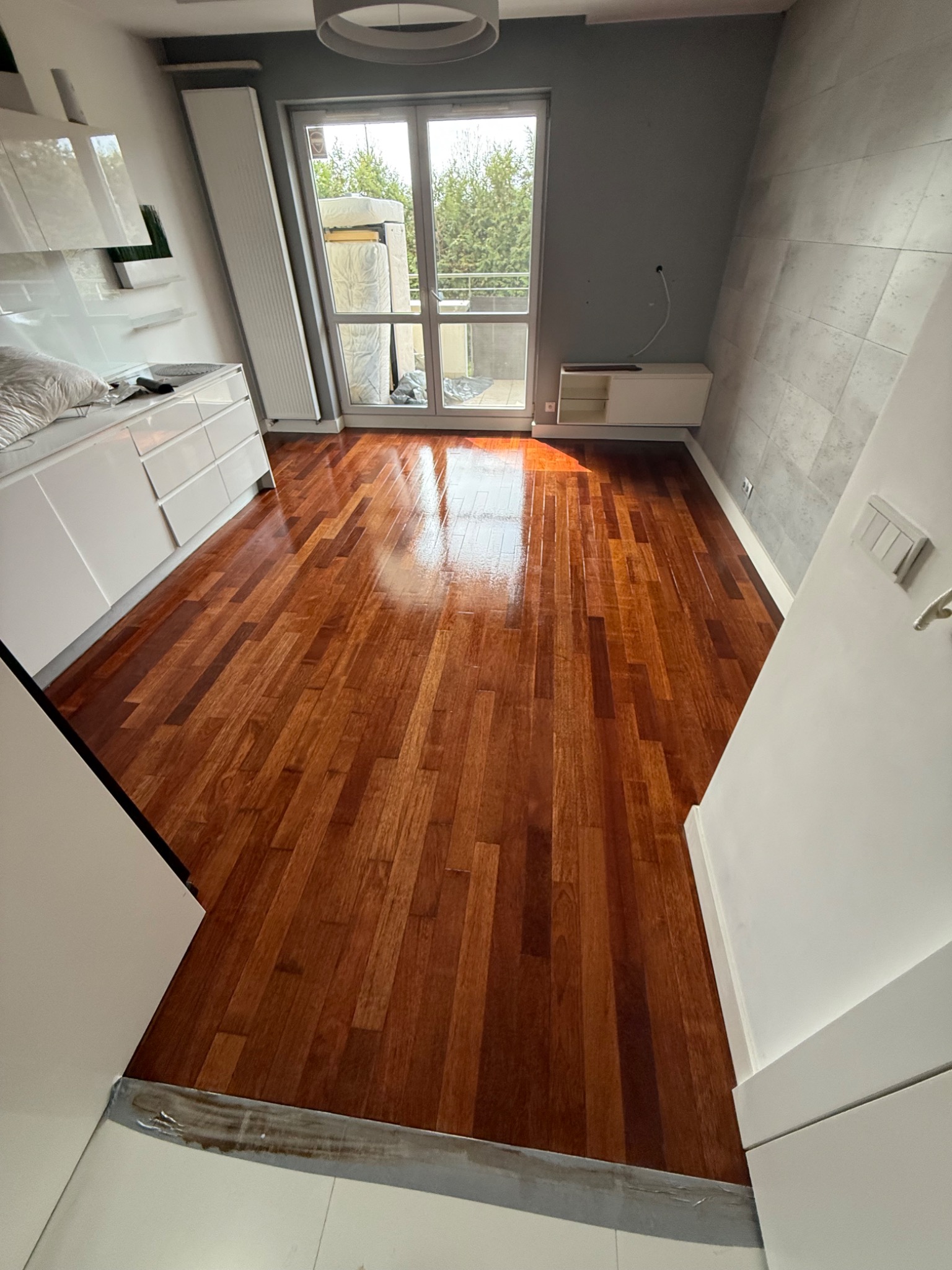 Błyszcząca, świeżo polakierowana podłoga parkietowa w jasnym pomieszczeniu z białymi meblami i szarą ścianą, widok z perspektywy wejścia.