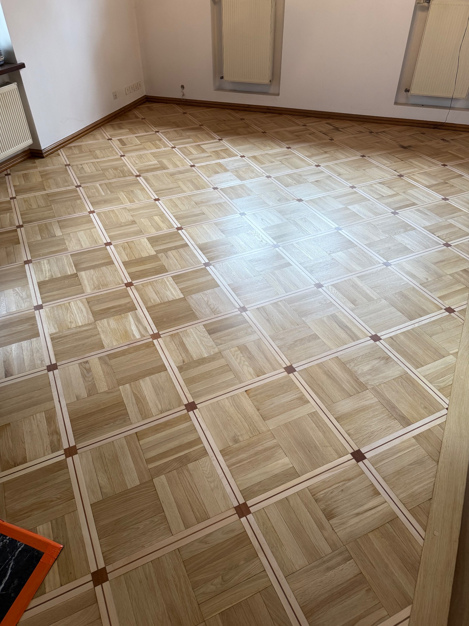 Ułożony parkiet w kwadraty z ciemniejszymi wstawkami, widoczne ślady po cyklinowaniu na niektórych fragmentach, w rogu fragment pomarańczowej folii ochronnej.